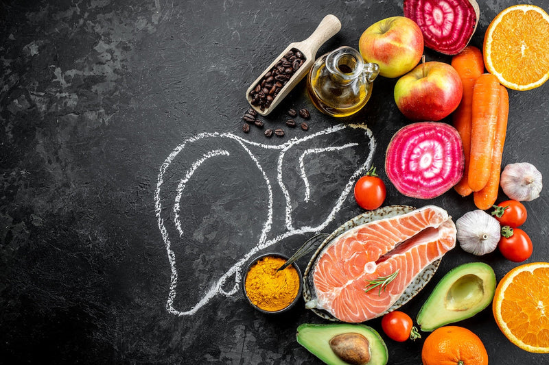 Chalk drawing of a liver surrounded by fresh fruits, vegetables, salmon, and other healthy foods on a dark surface.