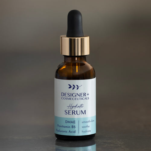 Designer+ Cosmeceuticals Hydrate Serum with DMAE, Provitamin B5, and hyaluronic acid in a brown glass dropper bottle, on a reflective surface with a dark background