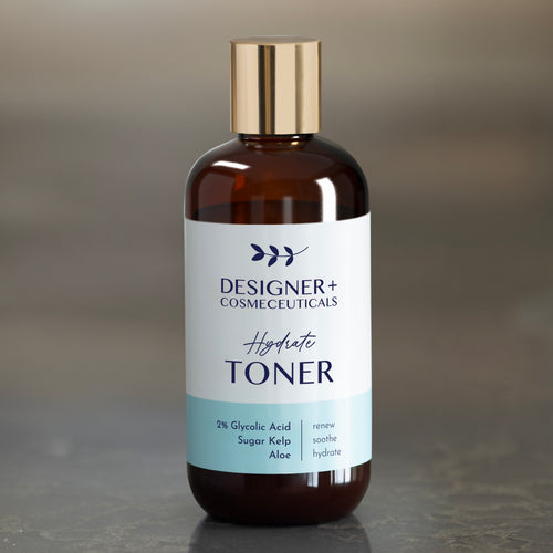 Designer+ Cosmeceuticals Hydrate Toner with 2% glycolic acid, sugar kelp, and aloe in a brown glass bottle with a gold cap, on a reflective surface with a gray background