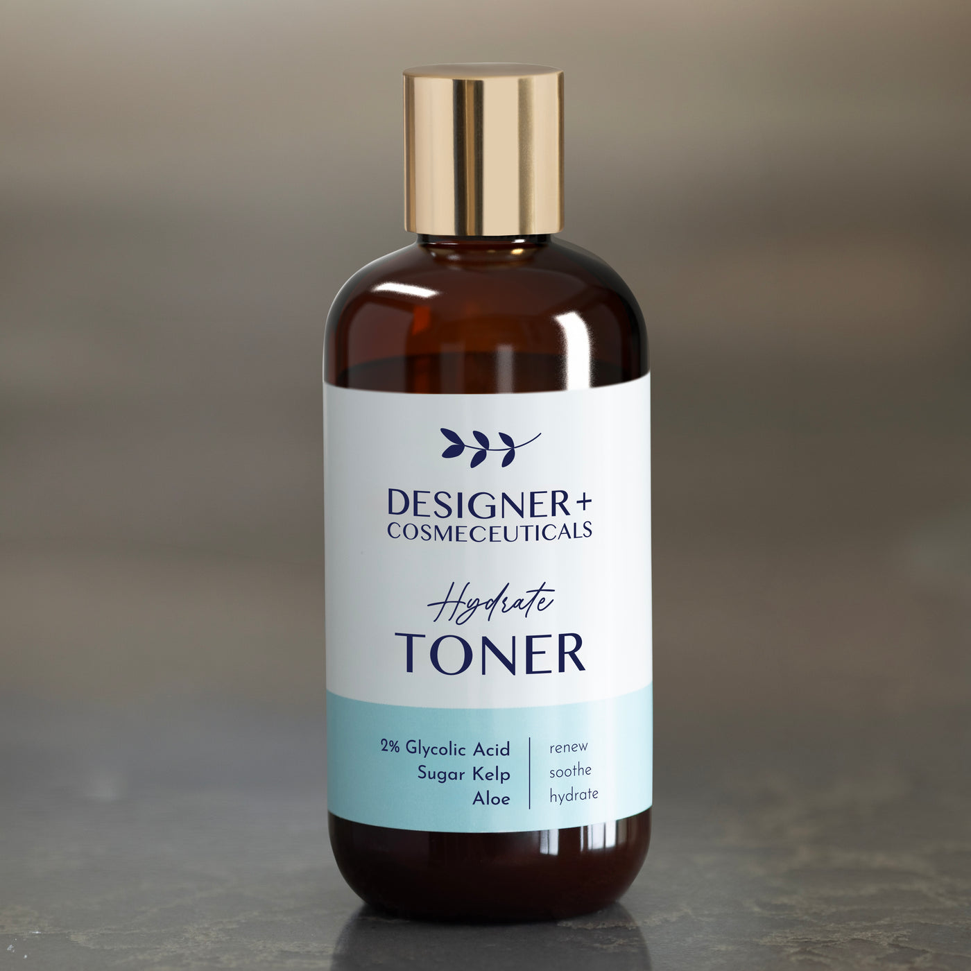 Designer+ Cosmeceuticals Hydrate Toner with 2% glycolic acid, sugar kelp, and aloe in a brown glass bottle with a gold cap, on a reflective surface with a gray background