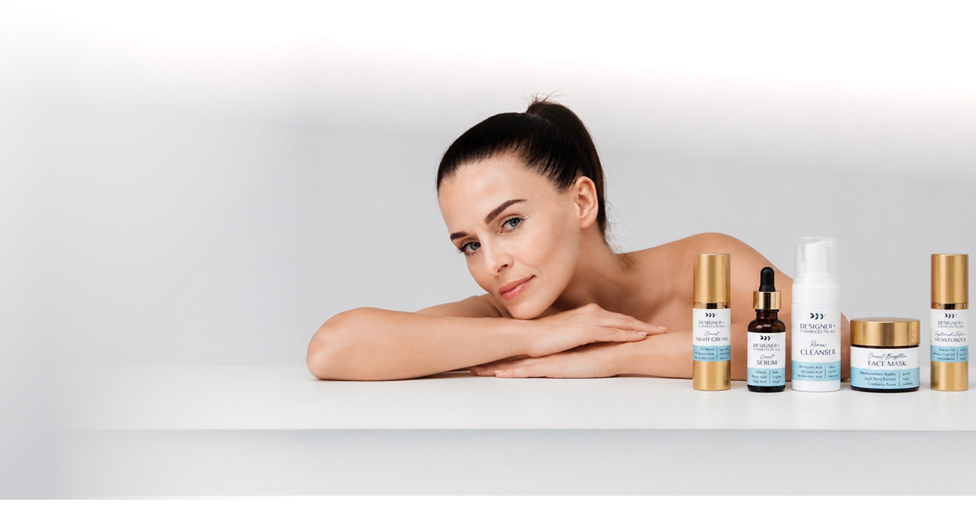 Woman with clear skin resting her chin on her arms beside a lineup of Designer+ skincare products including serum, cleanser, face mask, and cream, on a white background